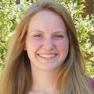 Rebecca Faust smiling, with long light brown hair, in front of green shrubs.